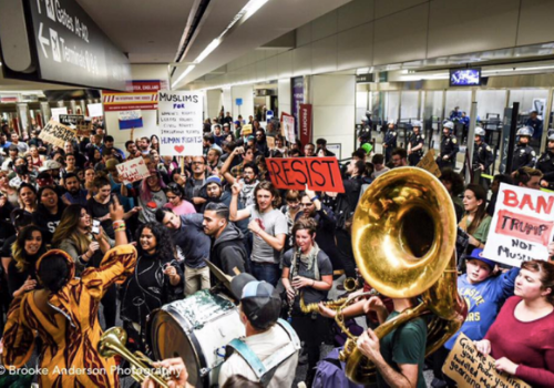 A group of people protesting at the San Francisco National Airport, holding signs that say "RESIST and "Ban Trump not Muslims" and holding instruments.